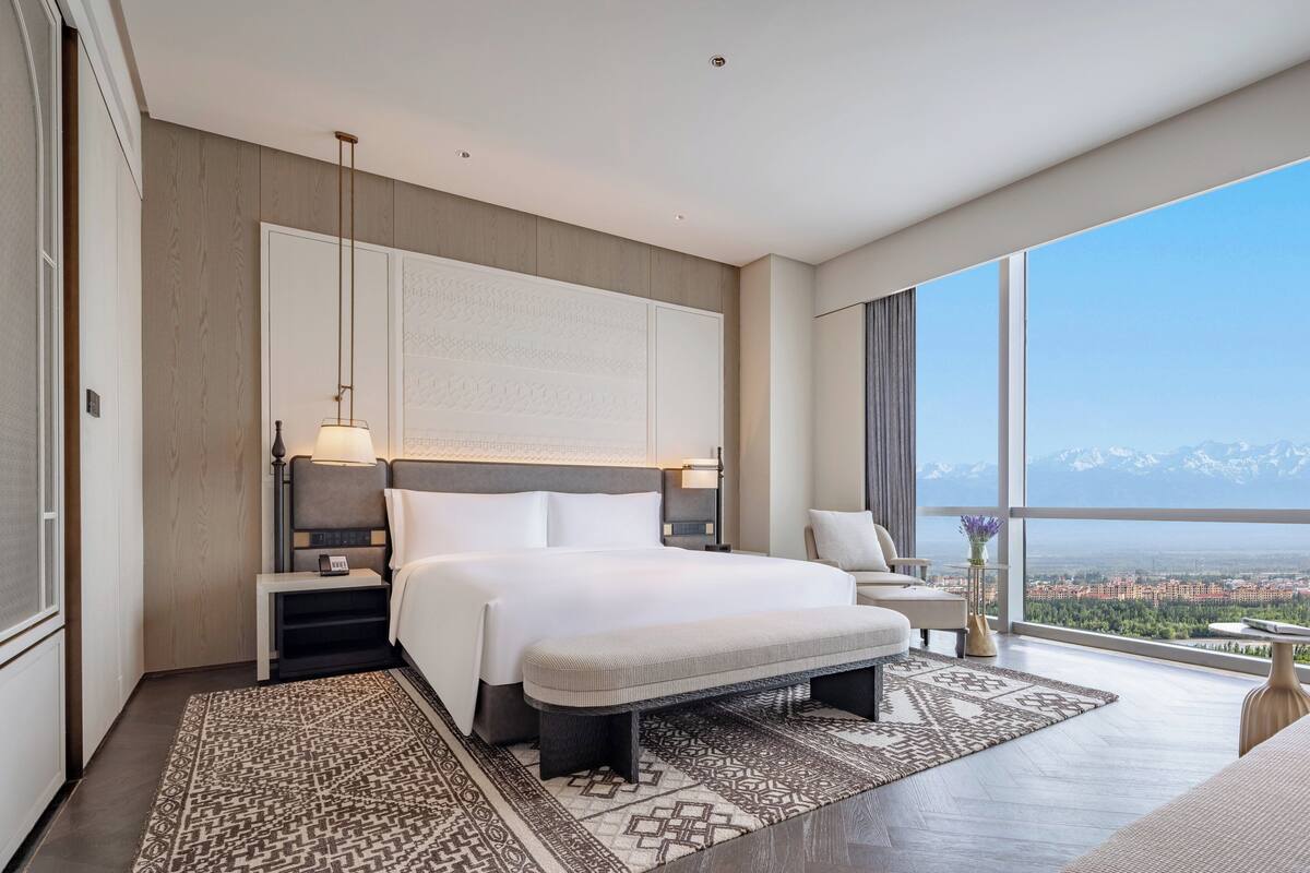 A modern hotel room with a large bed, a nightstand, a bedside table, a bench, and a view of a city and mountains.