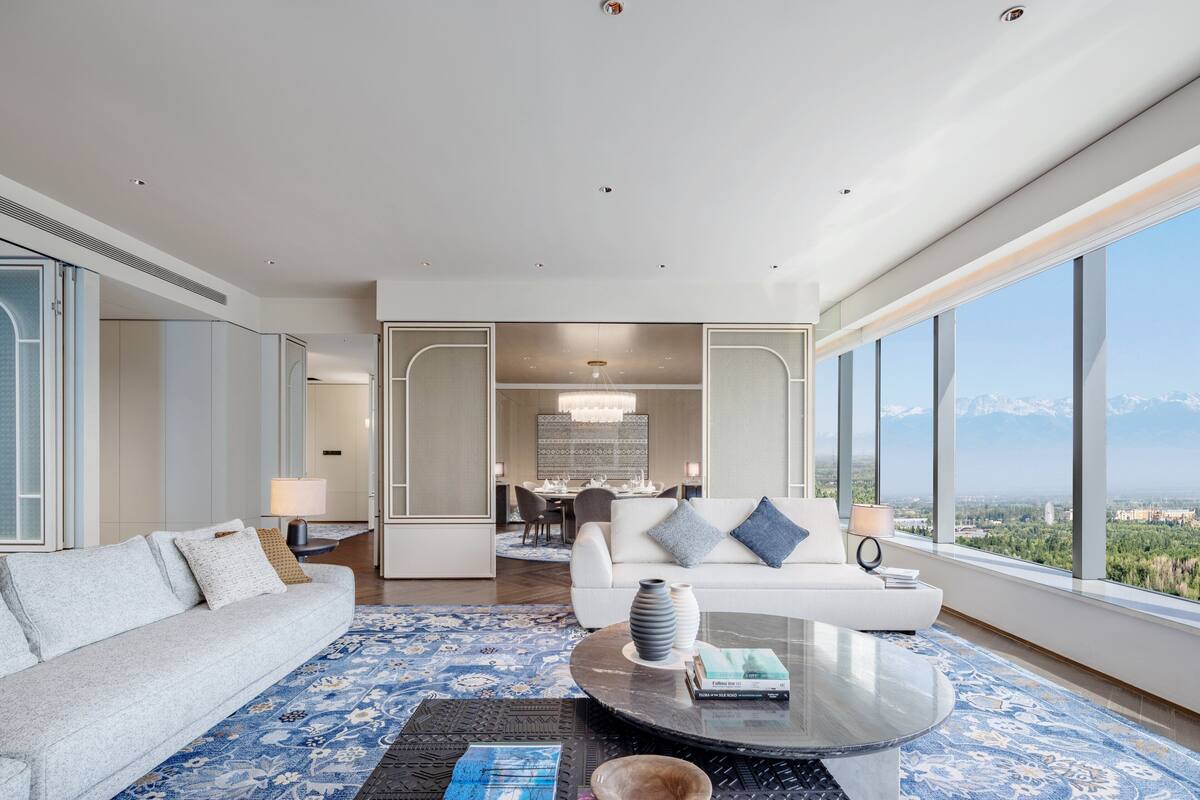 A modern hotel room with a large window offering a mountain view, a sofa, a coffee table, and a dining area in the background.