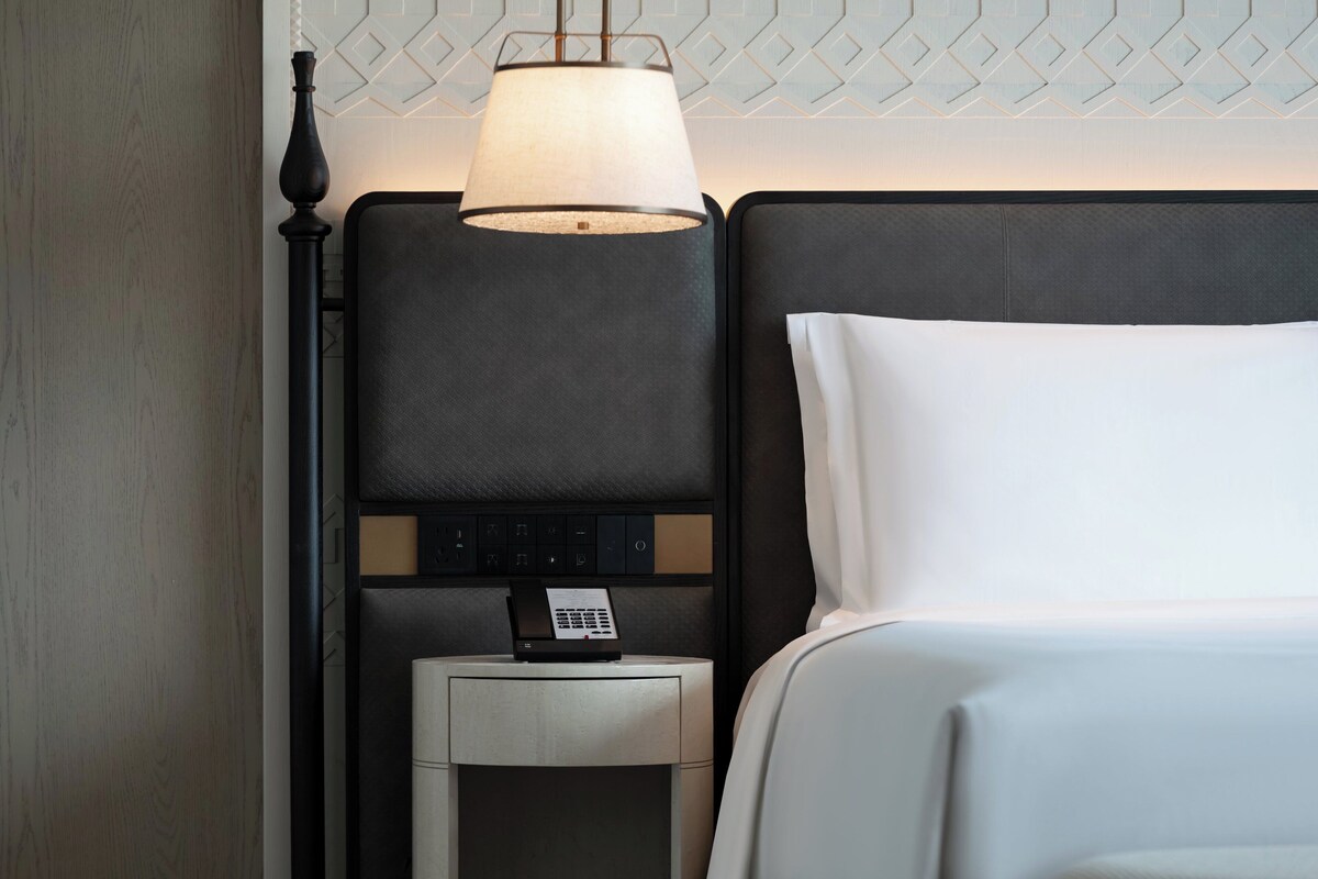 A neatly made hotel room with a bed, a bedside table with a lamp, and a control panel on the wall.
