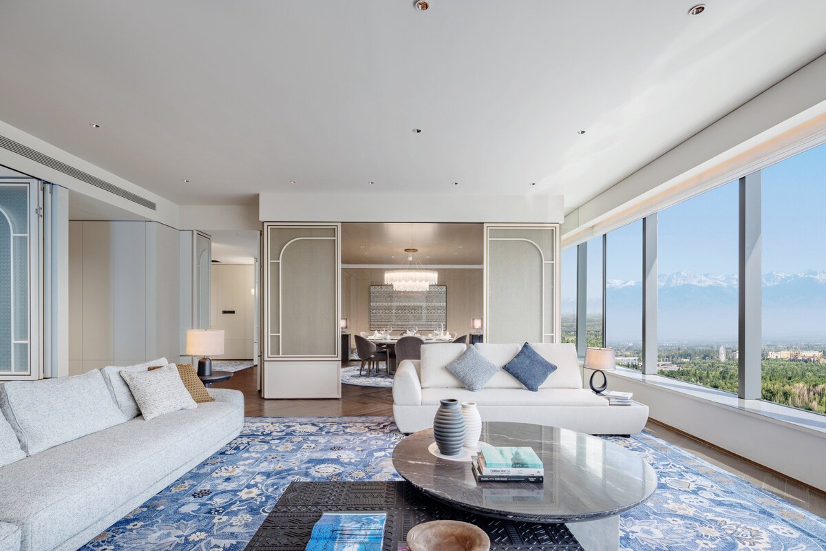 A modern hotel room with a large window offering a mountain view, a sofa, a coffee table, and a dining area in the background.