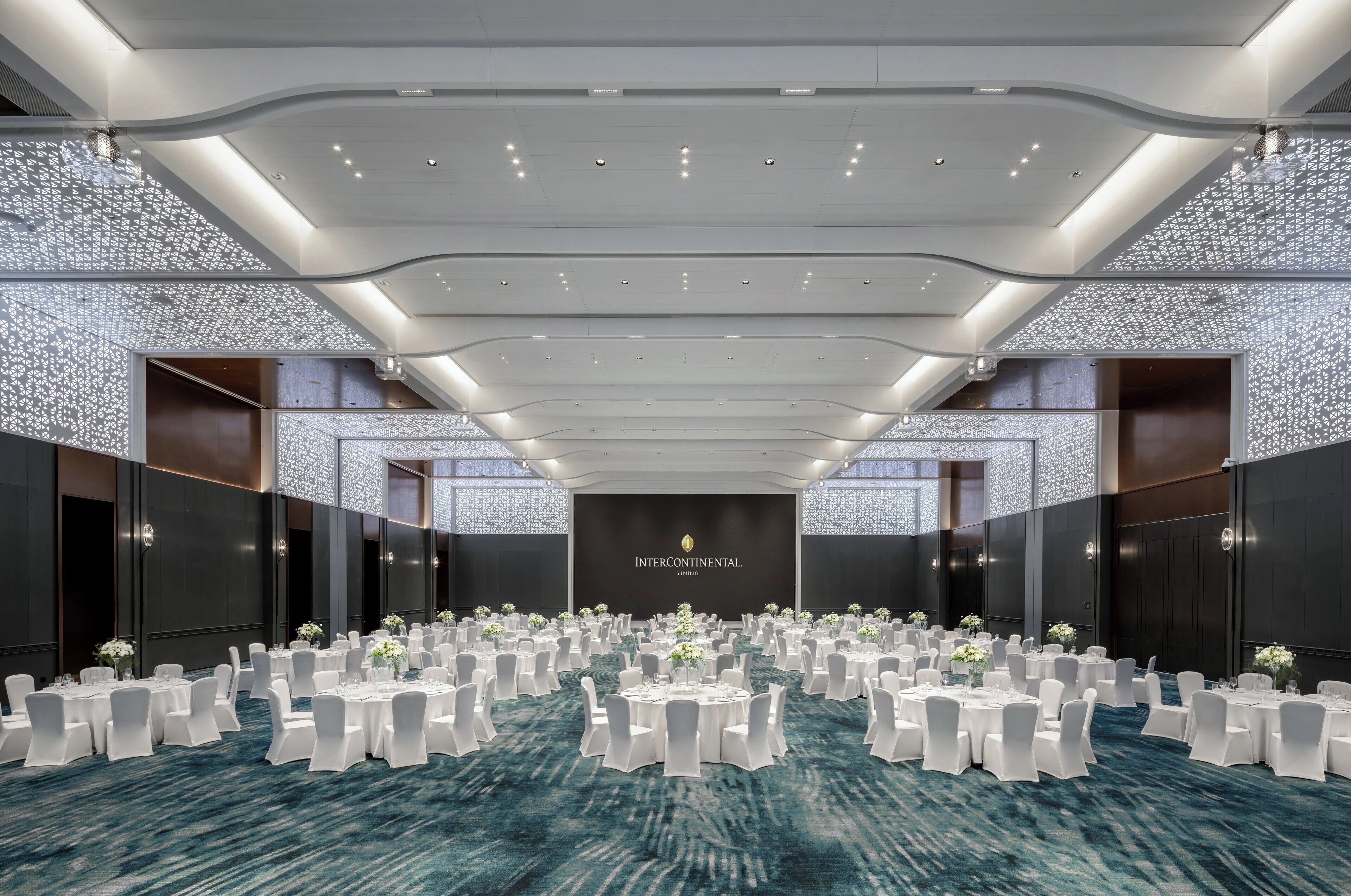 A spacious banquet hall with round tables and white chairs, a dark carpet, and a modern ceiling design.