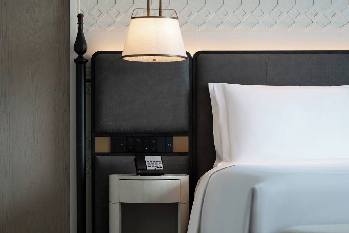 A neatly made hotel room with a bed, a bedside table with a lamp, and a control panel on the wall.