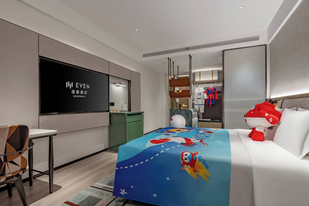 A modern hotel room with a large flat-screen TV, a neatly made bed with a space-themed bedspread, a comfortable chair, and a small table.