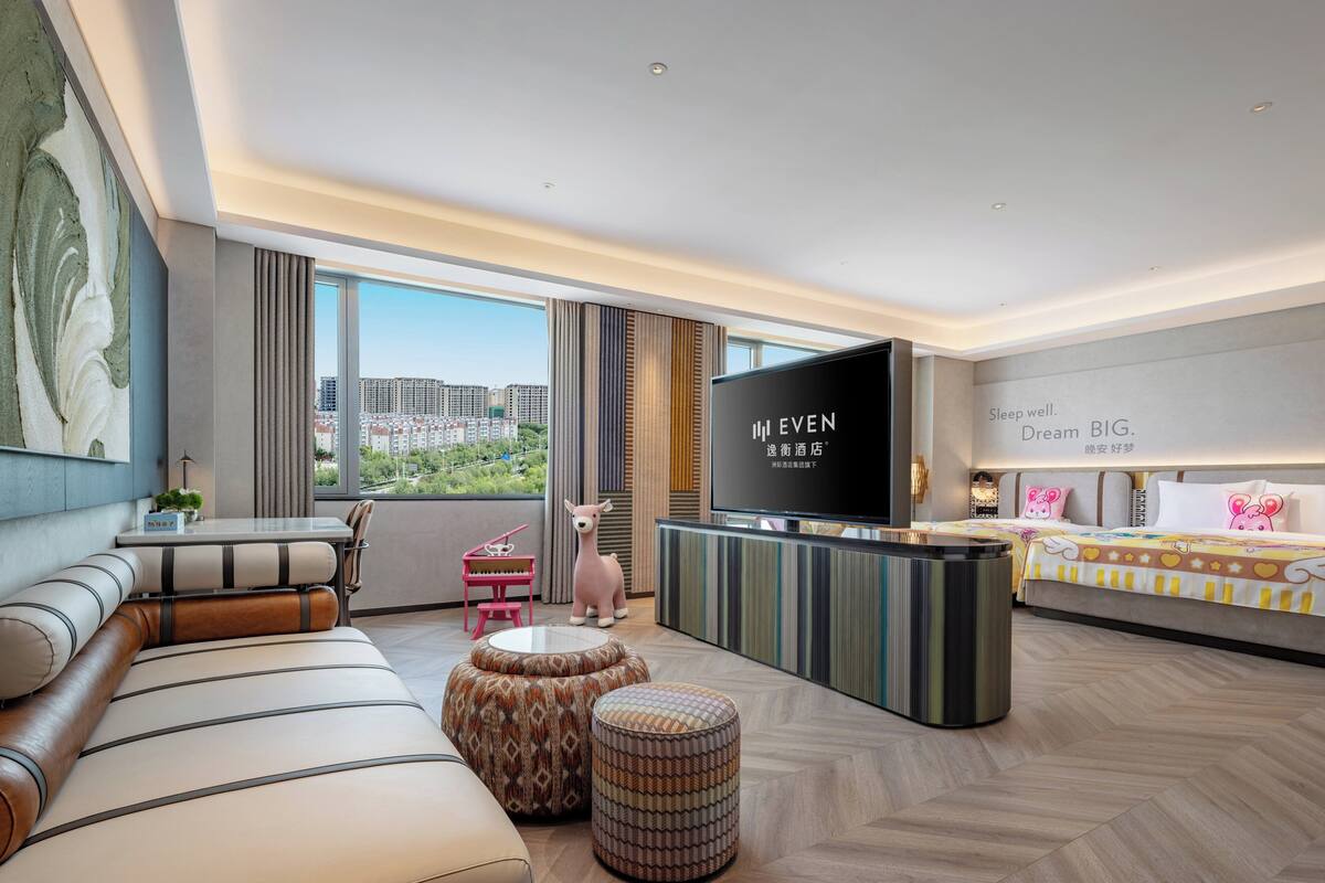 A modern hotel room with a large TV, a sofa, ottomans, a small pink chair, and a bed with a colorful headboard.