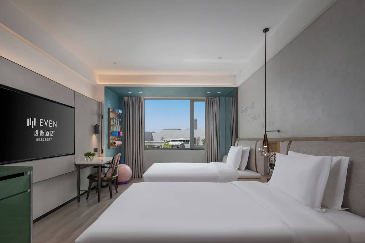 A hotel room with a large flat-screen TV, two beds, a desk with a chair, and a view of the cityscape.