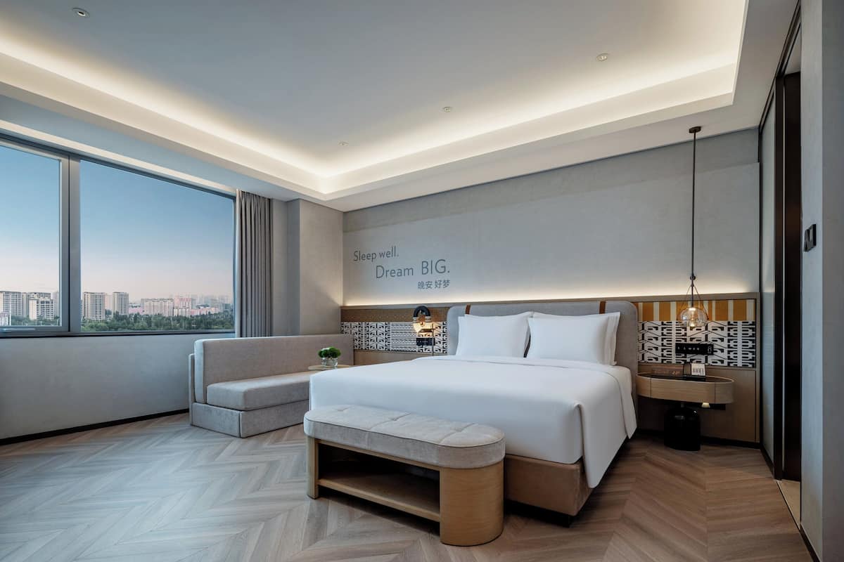 A modern hotel room with a large bed, a bench, a nightstand, and a view of the city.