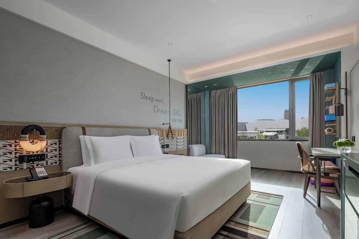 A modern hotel room with a large bed, a desk, and a view of the city.
