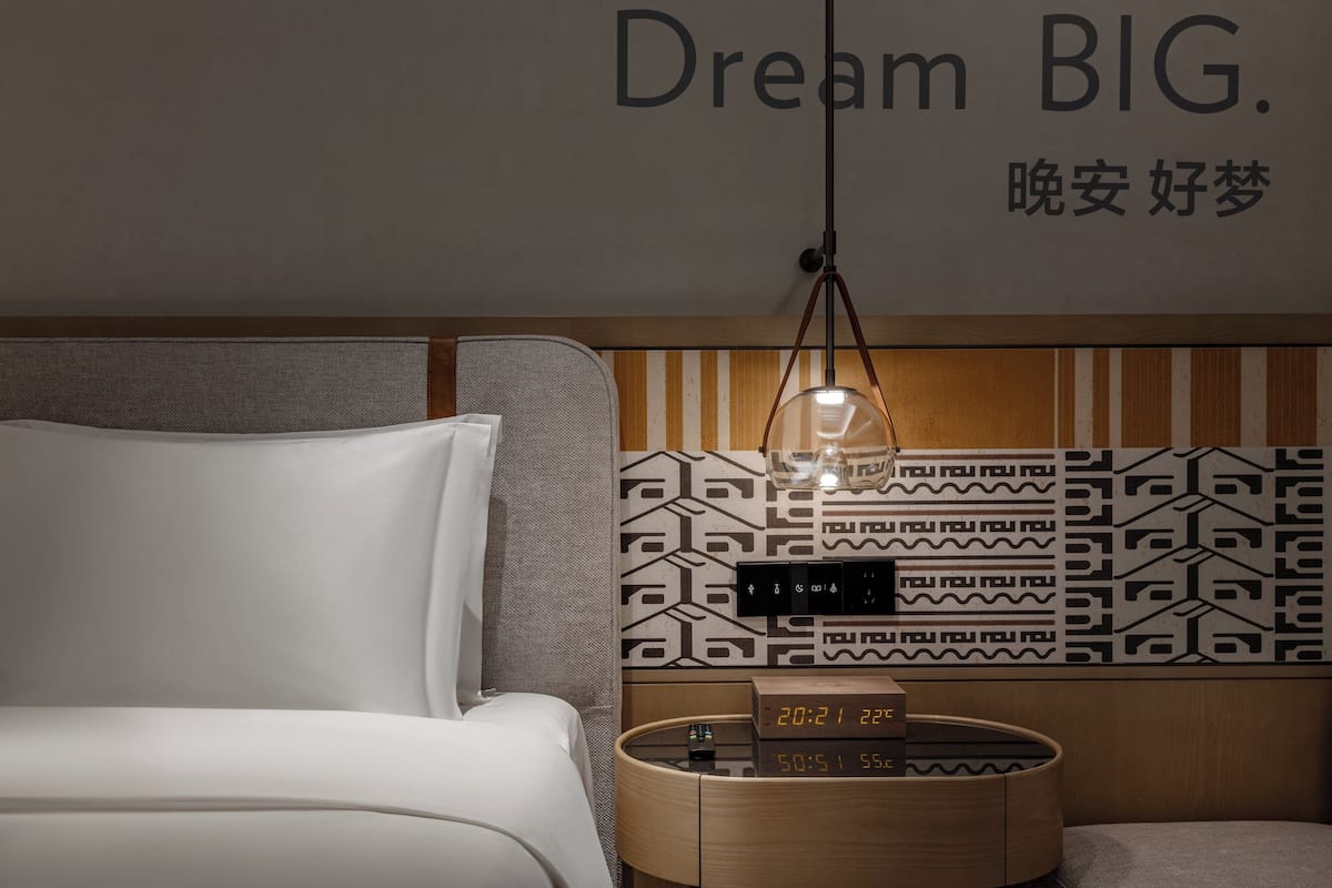 A modern hotel room with a bed, a bedside table with a clock, and a wall with a geometric pattern and the text "Dream BIG."