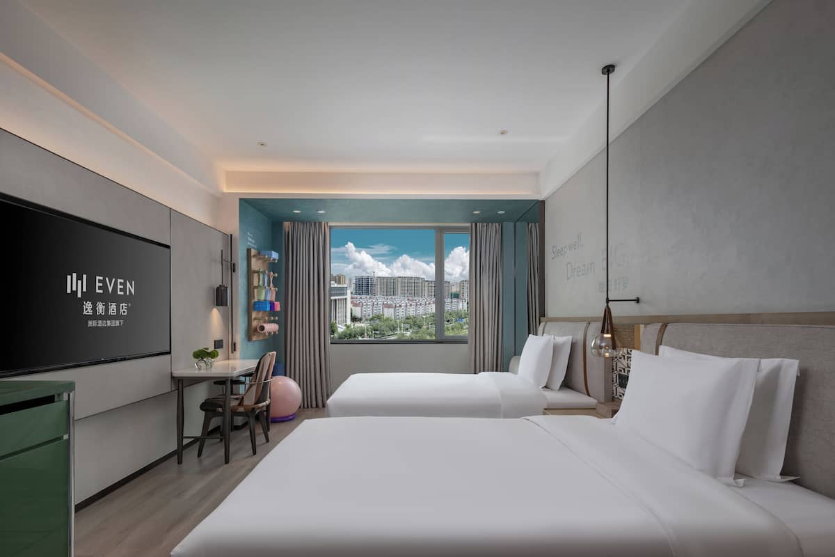 A modern hotel room with a large TV, a desk with a chair, two beds, and a city view.