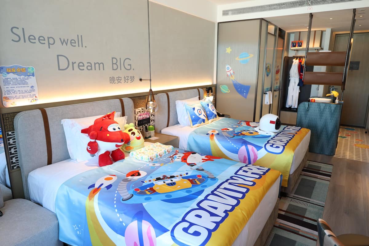 A hotel room with two beds, each with a Gravity-themed bedspread, a wall with a motivational message, and a children's play area visible through a glass partition.