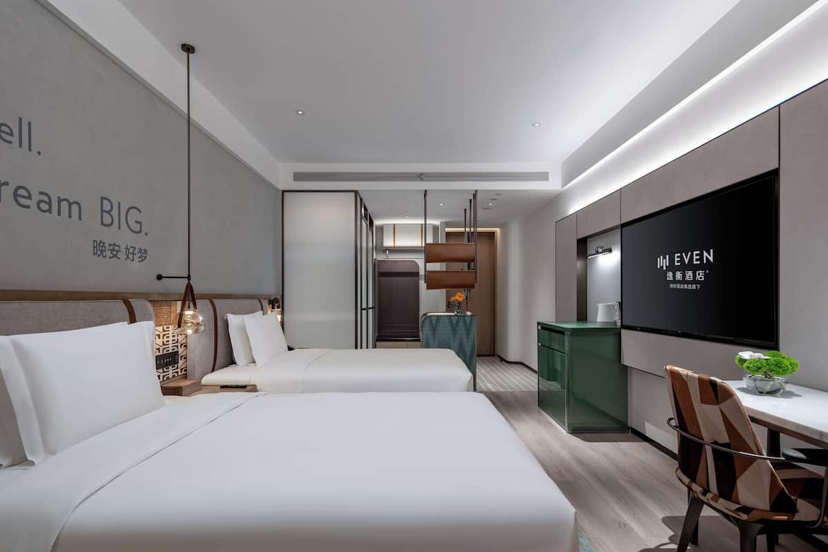 A modern hotel room with a large bed, a desk with a chair, a TV, and a small plant.