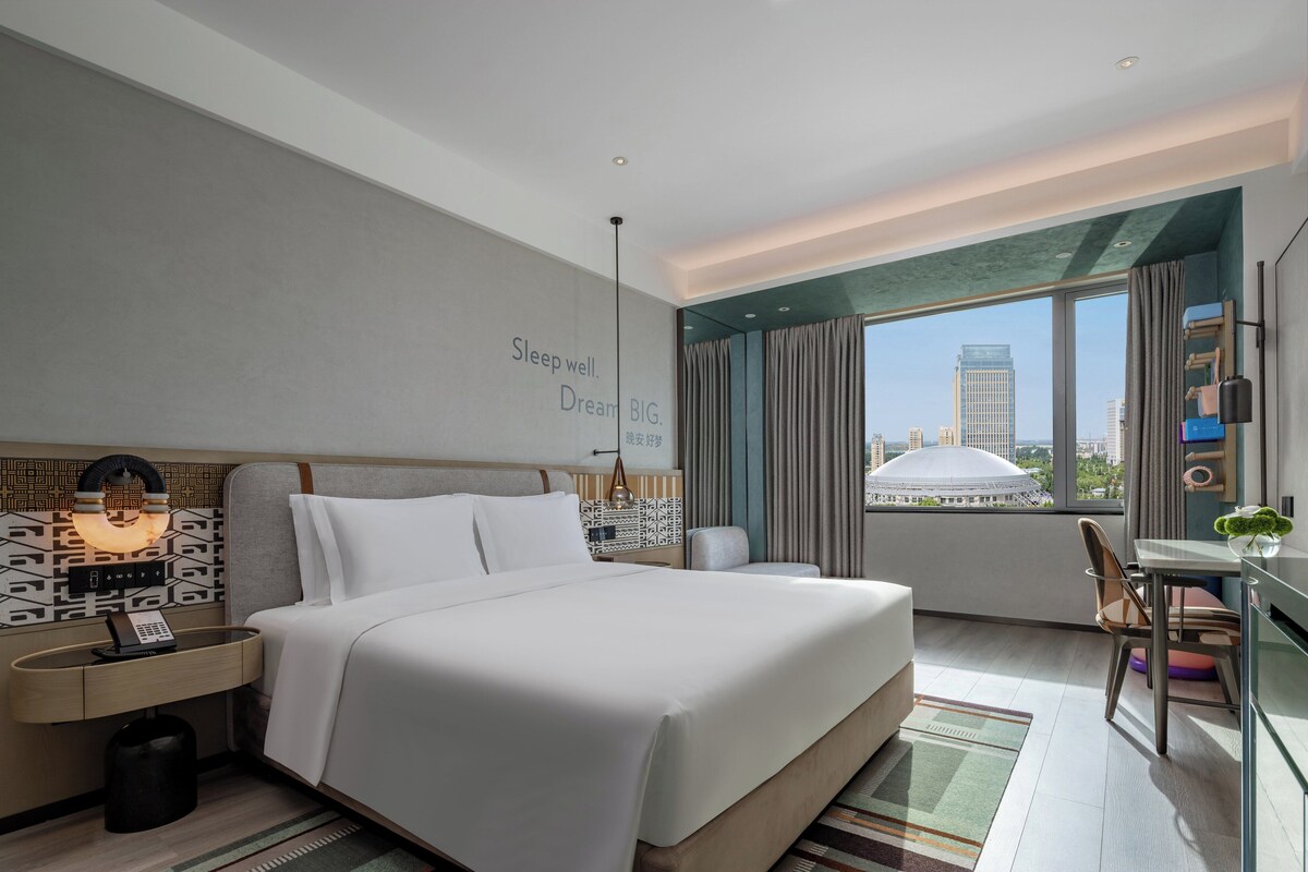 Une chambre d’hôtel moderne dotée d’un grand lit, d’un bureau avec une chaise, d’une vue sur le paysage urbain et d’un mur orné d’une citation inspirante.
