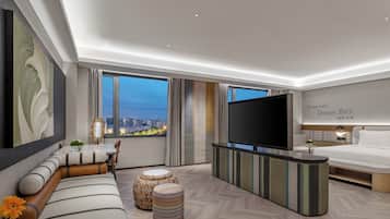 Quarto de hotel moderno com uma cama grande, televisão de tela plana, sofá, pufes e vista para a cidade.