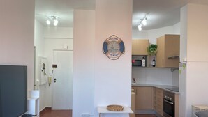 A modern living room with a flat-screen TV, a small table, and a sofa. There is a kitchen area with wooden cabinets and a microwave.