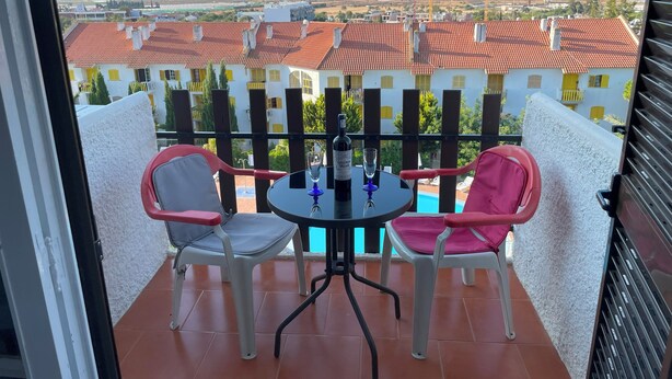 A balcony with two chairs and a table, overlooking a pool and buildings with red-tiled roofs.