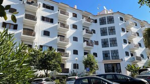 A multi-story building with balconies, parked cars, and a clear blue sky.
