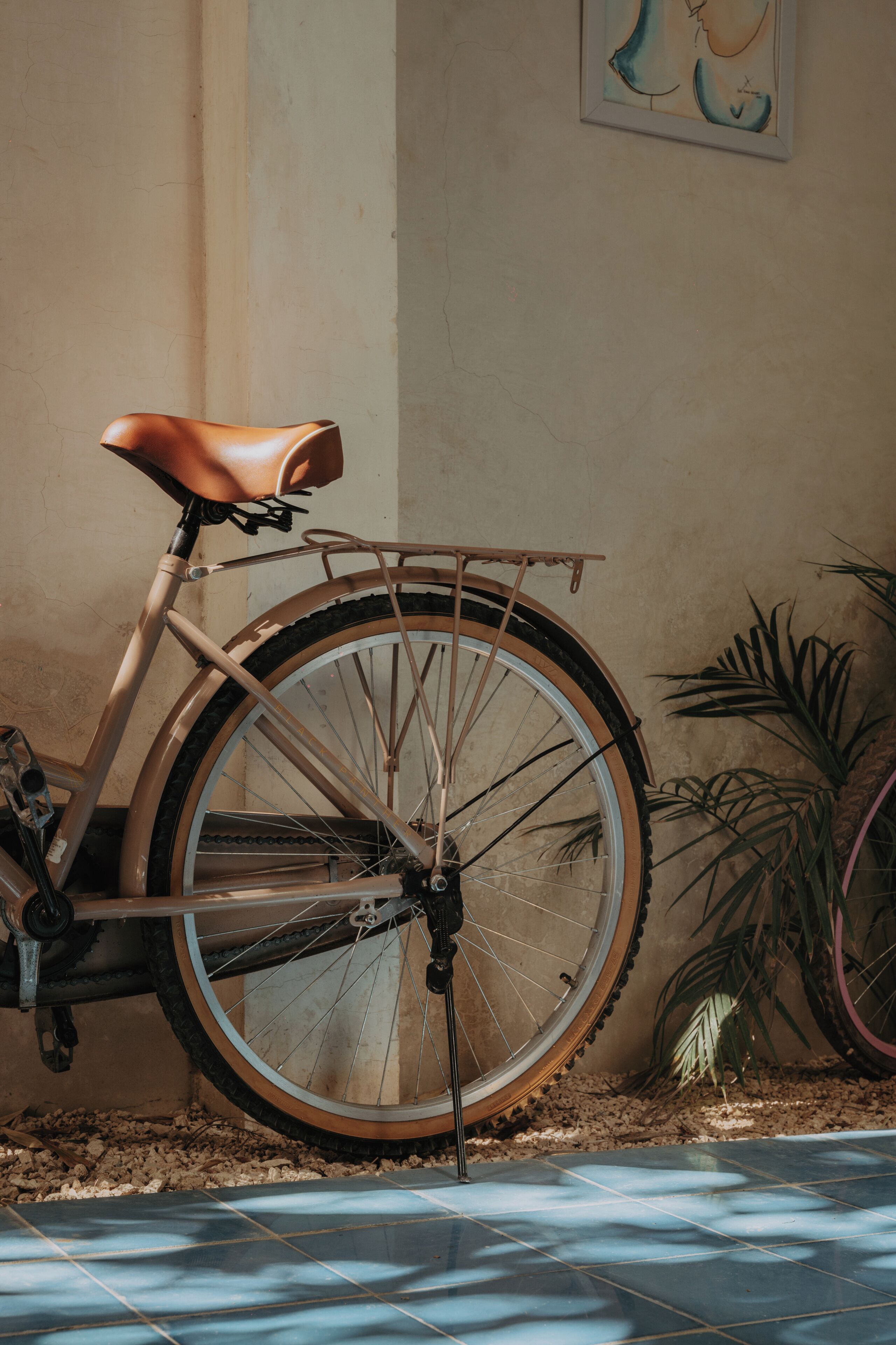 A bicycle with a leather seat and a front basket, parked indoors with a tiled floor and a plant in the background.