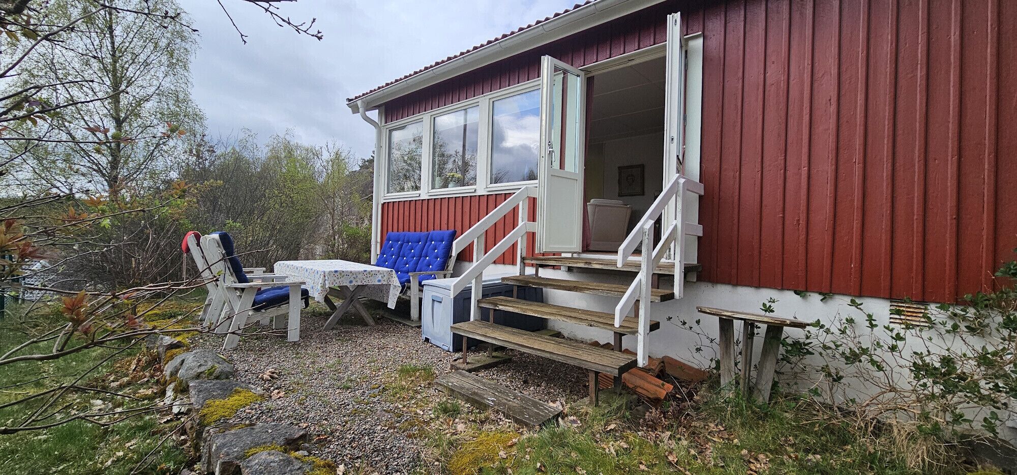 Een chalet met een rode gevel, een witte deur, een klein terras met een tafel en twee stoelen, en een trap die naar de ingang leidt.