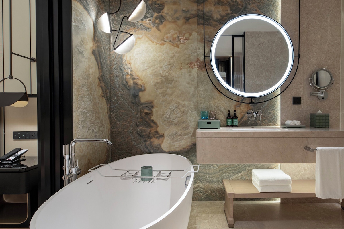 Een moderne badkamer met een vrijstaand bad, een ronde spiegel, een wastafelmeubel met een wastafel en een bankje met handdoeken.