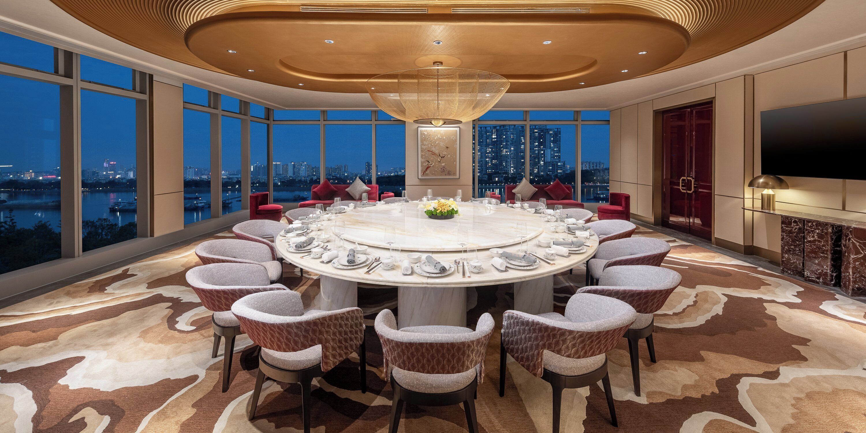 A high-rise hotel room with a round dining table set for eight, surrounded by plush chairs, a large chandelier, and a city view.