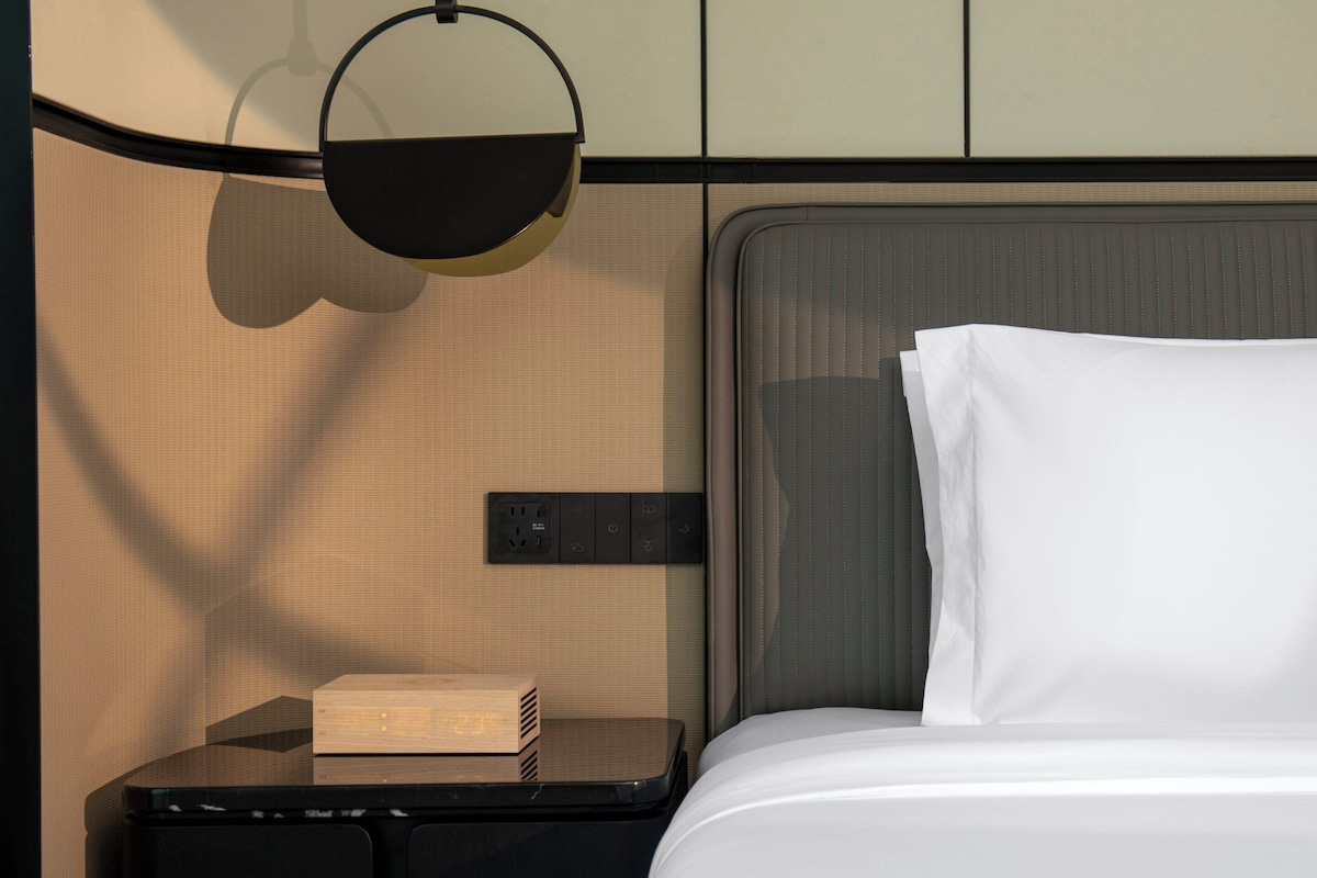 Een opgemaakt bed met witte lakens, een nachtkastje met een lamp, en een stopcontact op de muur.