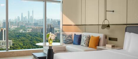 A modern hotel room with a city view, a sofa with patterned cushions, a bedside table, and a small bedside lamp.