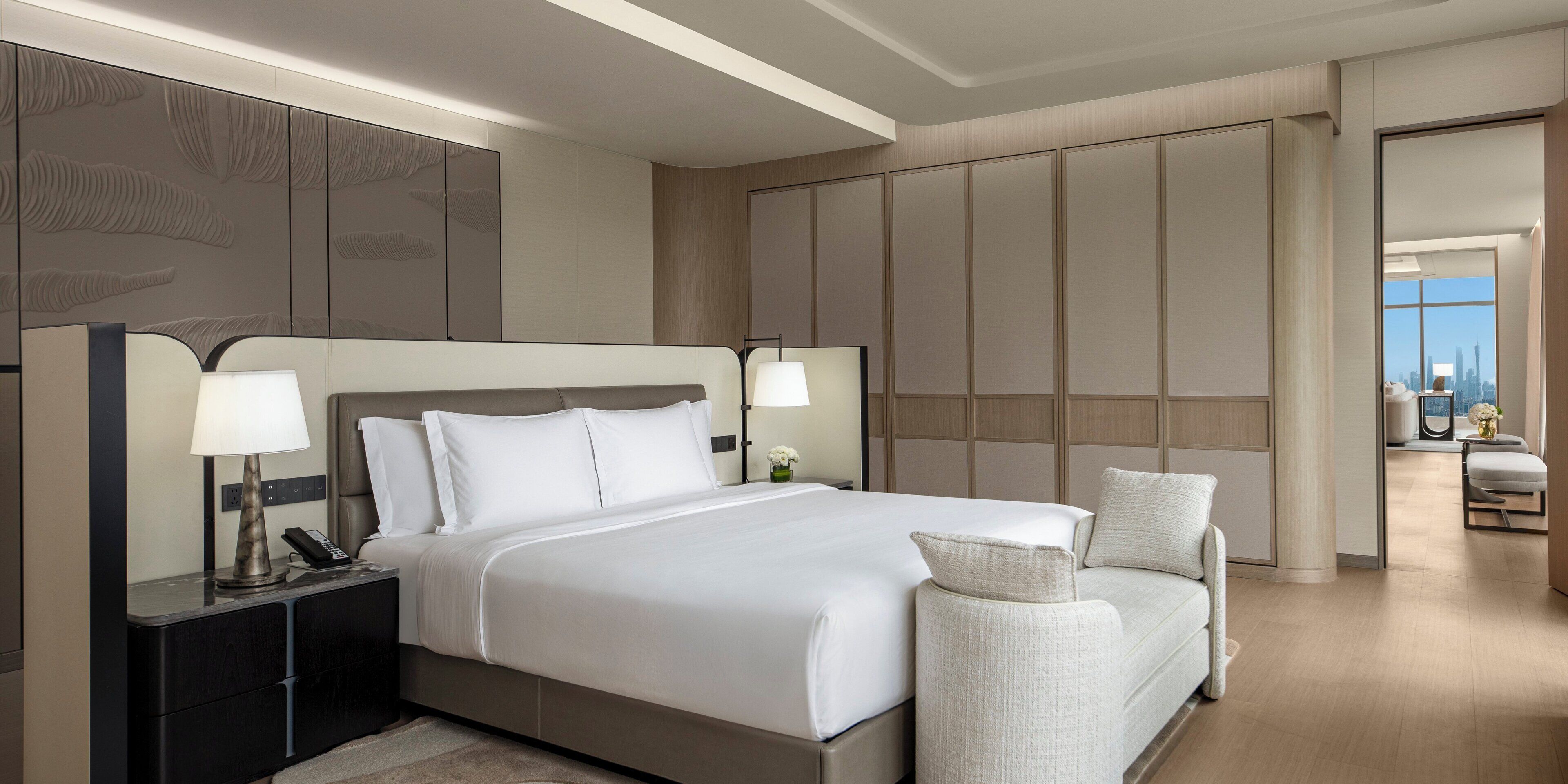 Ein modernes Hotelzimmer mit einem großen Bett, zwei Nachttischen mit Lampen, einer Sitzecke mit einem Sessel und Blick auf die Stadt durch große Fenster.