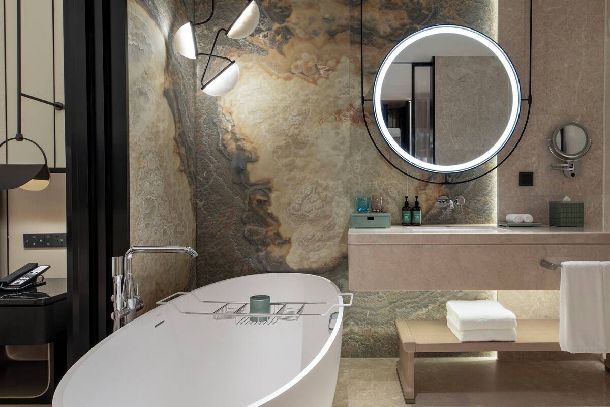 Ein modernes Badezimmer mit einer freistehenden Badewanne, einem runden Spiegel, einem Waschtisch und einer Sitzbank mit Handtüchern.