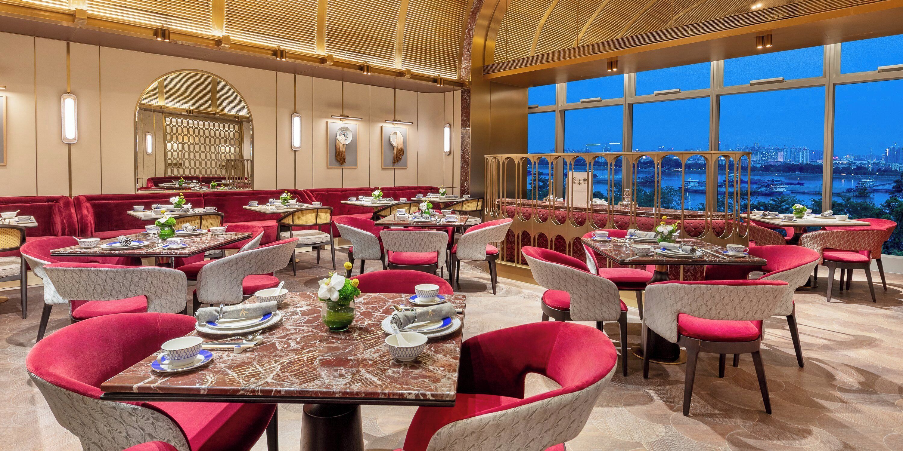 A restaurant with red upholstered chairs and tables, a large arched mirror, and a view of a cityscape through large windows.