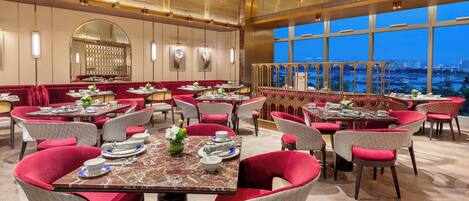 A restaurant with red upholstered chairs and tables, a large arched mirror, and a view of a cityscape through large windows.