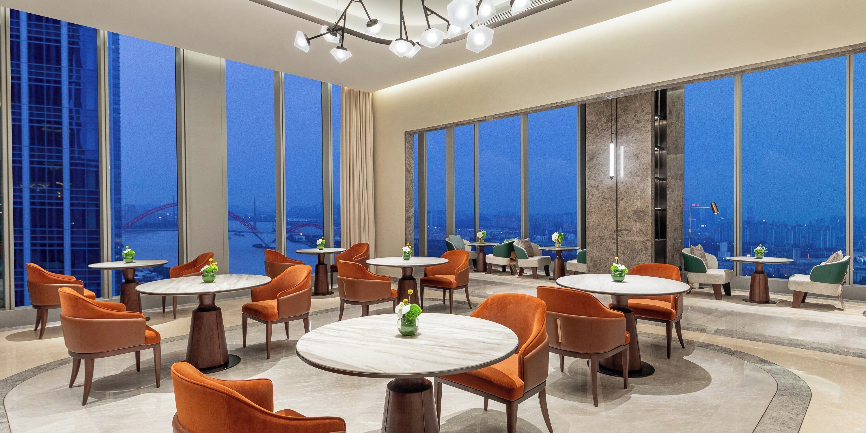 Una hall d'albergo moderna con tavoli rotondi e poltrone arancioni, ampie finestre con vista sulla città e un soffitto alto impreziosito da un lampadario originale.