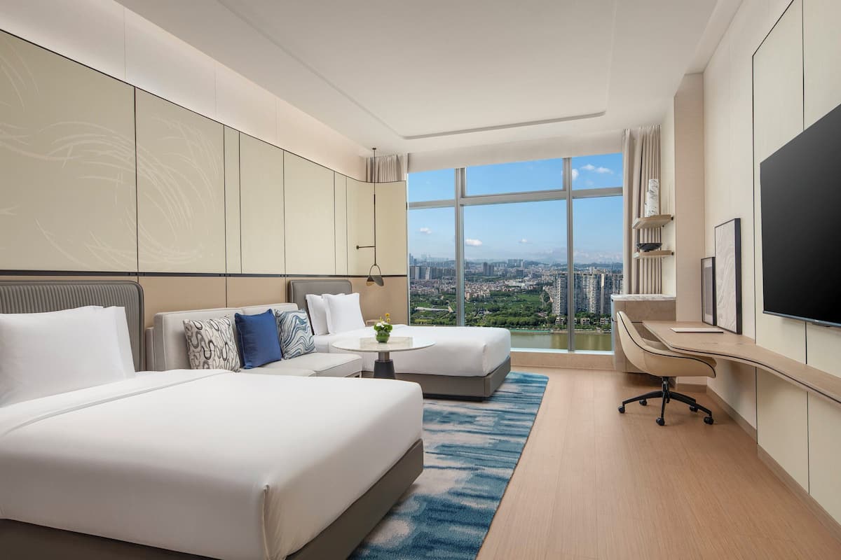 Ein Hotelzimmer mit einem großen Bett, einem Einzelbett, einem Schreibtisch und einem Fernseher. Große Fenster bieten Blick auf die Stadt.