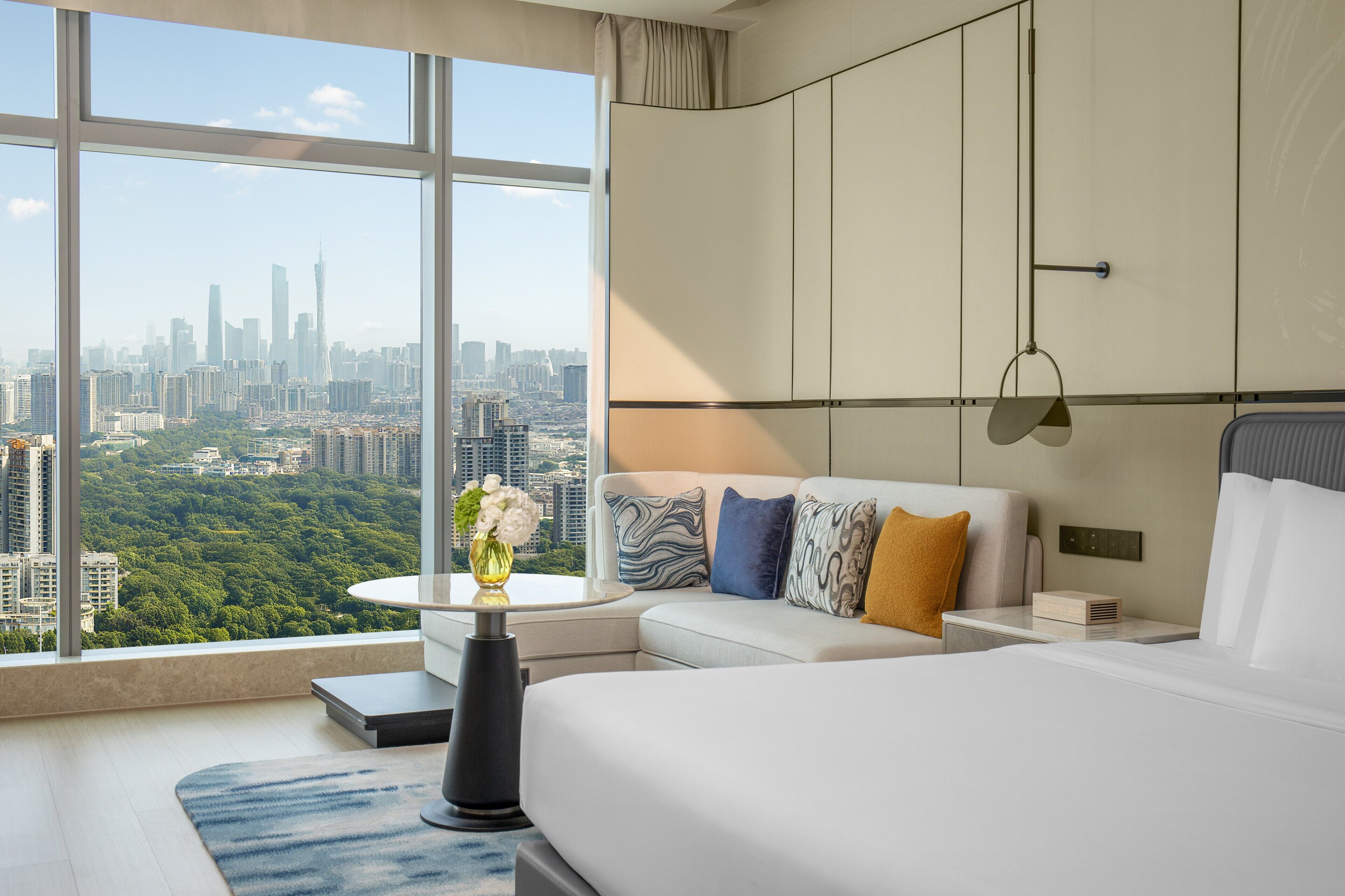 A modern hotel room with a large window offering a city view, a bed, a sofa, a small table with a vase, and a nightstand with a lamp.