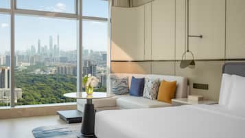 A modern hotel room with a large window offering a city view, a bed, a sofa, a small table with a vase, and a nightstand with a lamp.