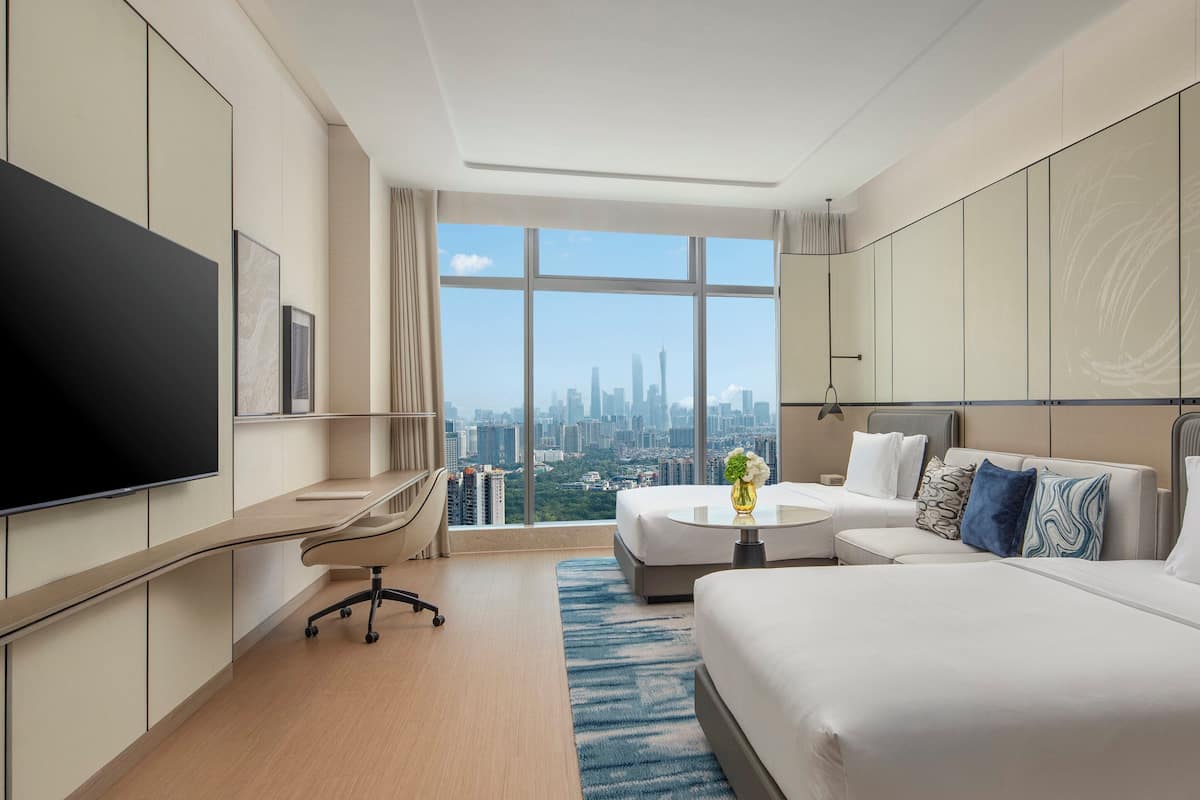Ein modernes Hotelzimmer mit einem großen Bett, einem Schreibtisch und einem Fernseher. Durch große Fenster bietet das Zimmer Blick auf die Stadt.