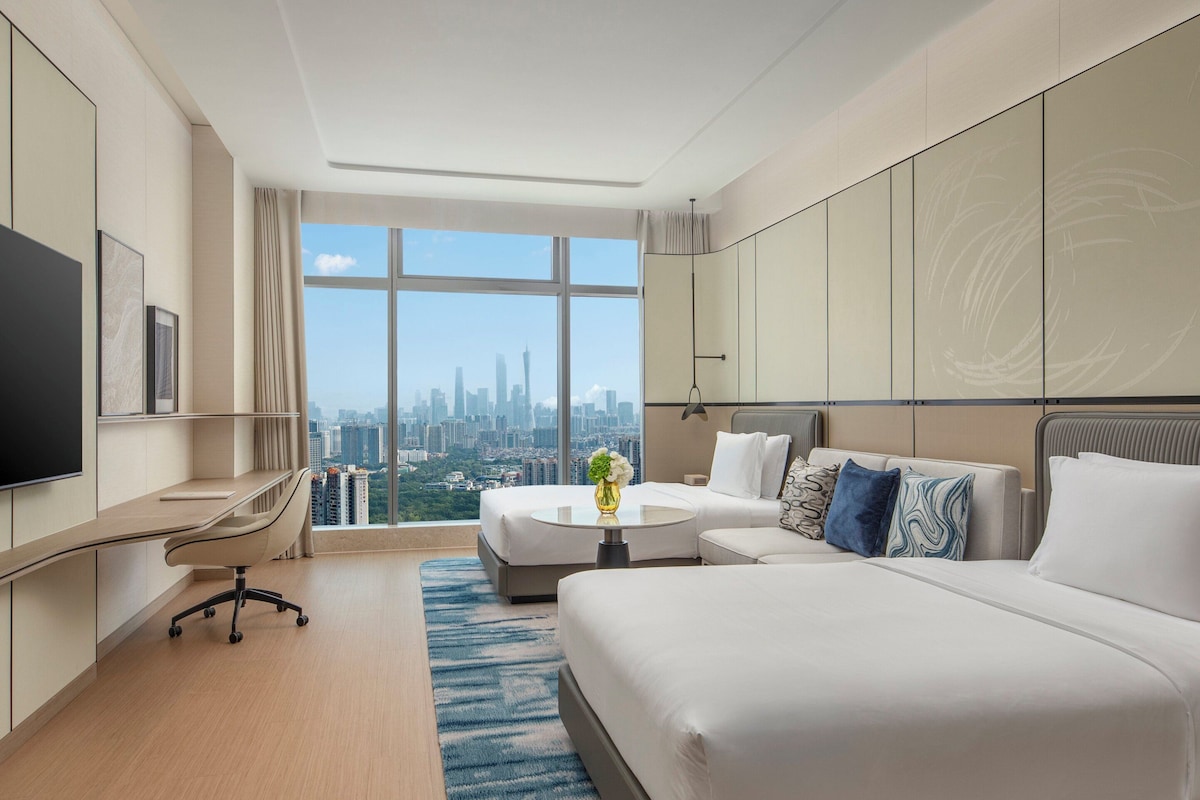 Een moderne hotelkamer met een groot bed, een bureau en een televisie. Door grote ramen is er uitzicht op de stad.