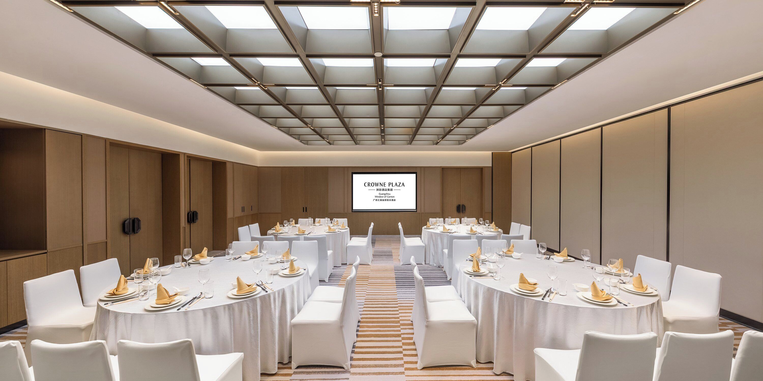 Un salón de conferencias con mesas redondas, sillas blancas y una pantalla grande que muestra 'CROWN PLAZA'.