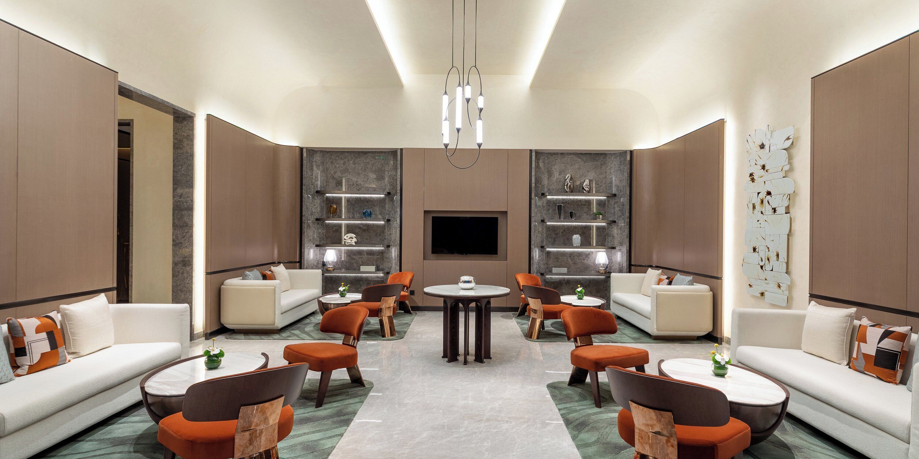 A modern hotel lobby with a central seating area, sofas, and armchairs, a fireplace, and a large wall-mounted TV.