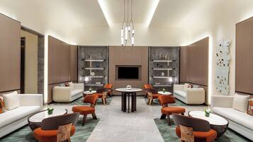 A modern hotel lobby with a central seating area, sofas, and armchairs, a fireplace, and a large wall-mounted TV.