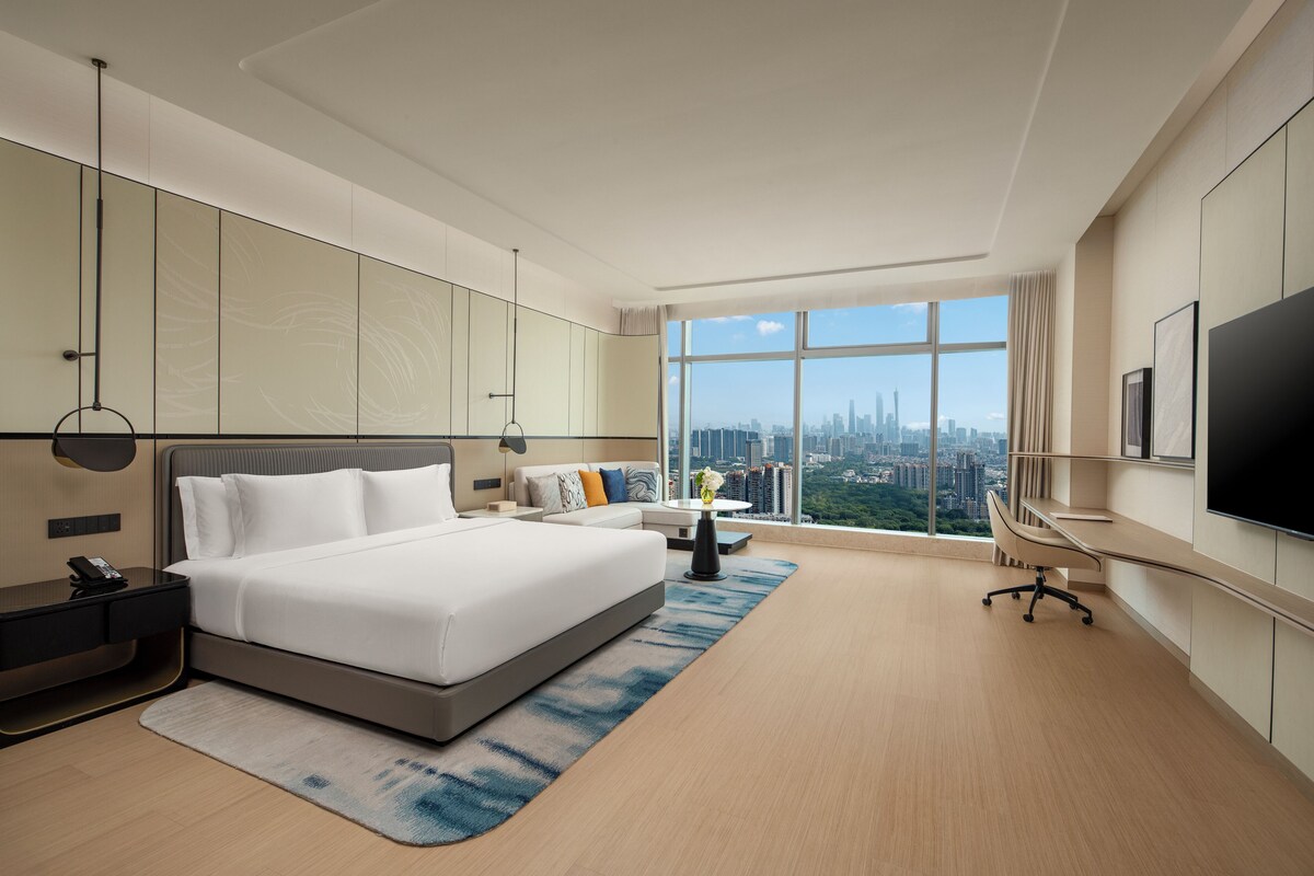 A modern hotel room with a large bed, a desk, and a view of the city skyline.