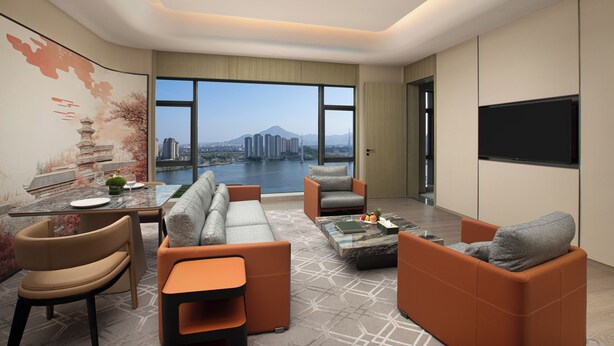 A modern living room with a sofa, armchairs, a coffee table, and a dining area. A large window offers a view of a cityscape and a body of water. - Crowne Plaza Putian by IHG (Putian)