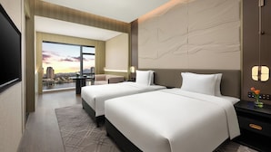 Habitación de hotel con dos camas, un ventanal grande con vista a la ciudad, un diseño moderno en la pared y una mesita de noche con un florero.