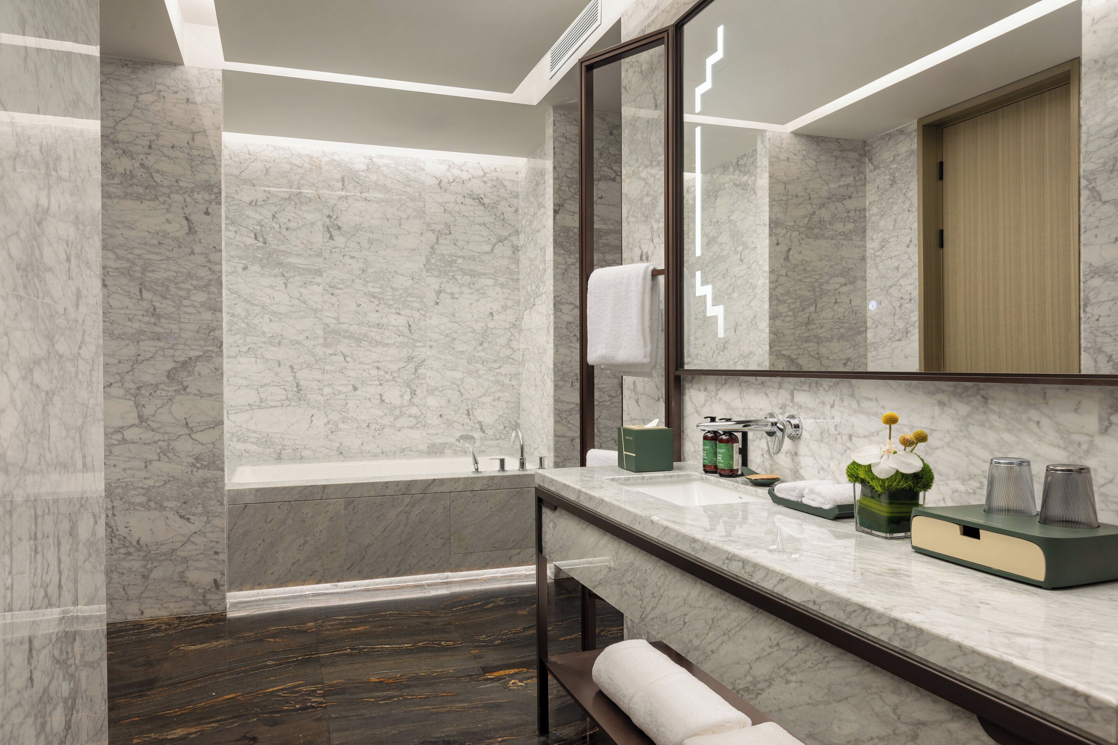A modern bathroom with a marble wall, a bathtub, a vanity with a mirror, and a wooden door.