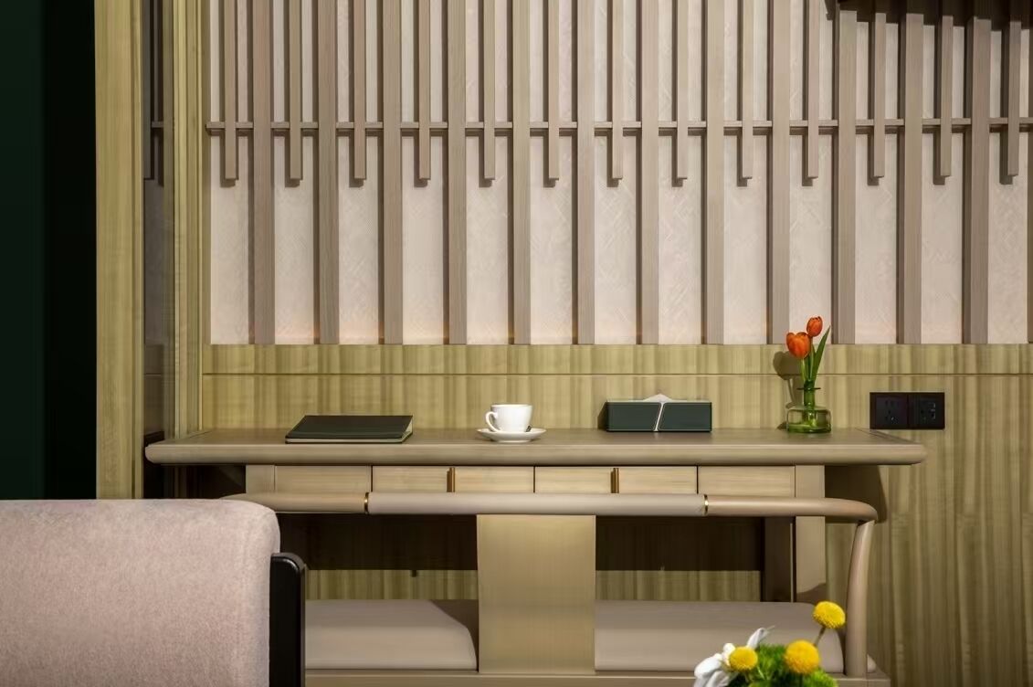 A modern hotel room with a minimalist design, featuring a long, low-profile desk with a tablet, a cup, and a vase with flowers.