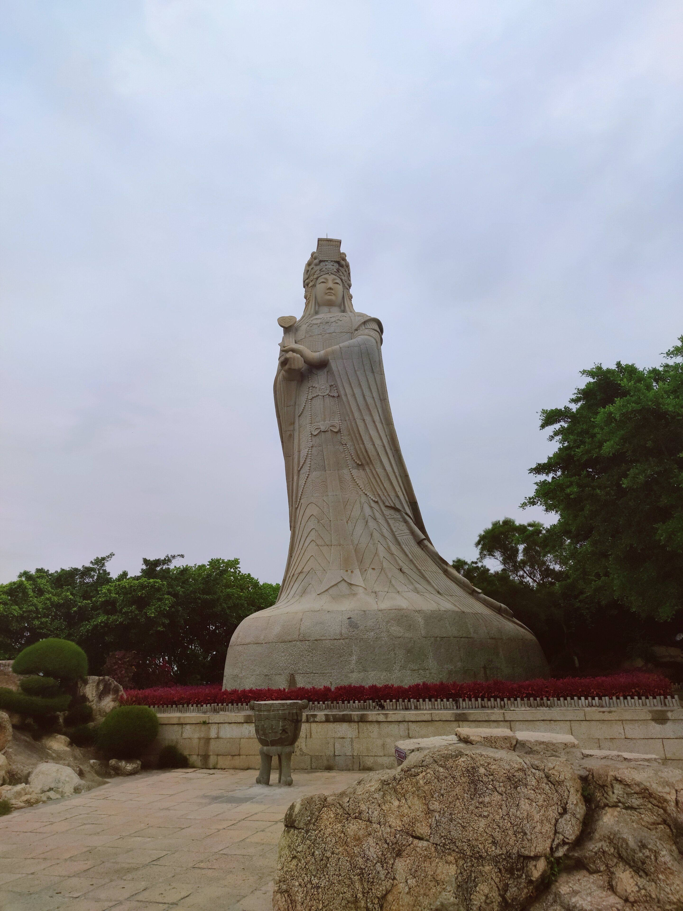 Una gran estatua de piedra que representa una figura vestida con traje tradicional, sobre un pedestal de piedra, con un jardín circundante y una urna grande al frente.