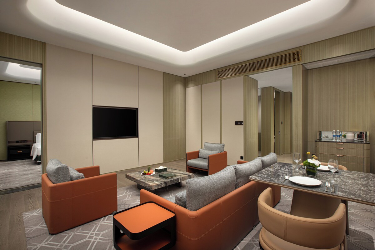 A modern hotel room with a seating area, a flat-screen TV, a dining table with a vase of flowers, and a minibar.