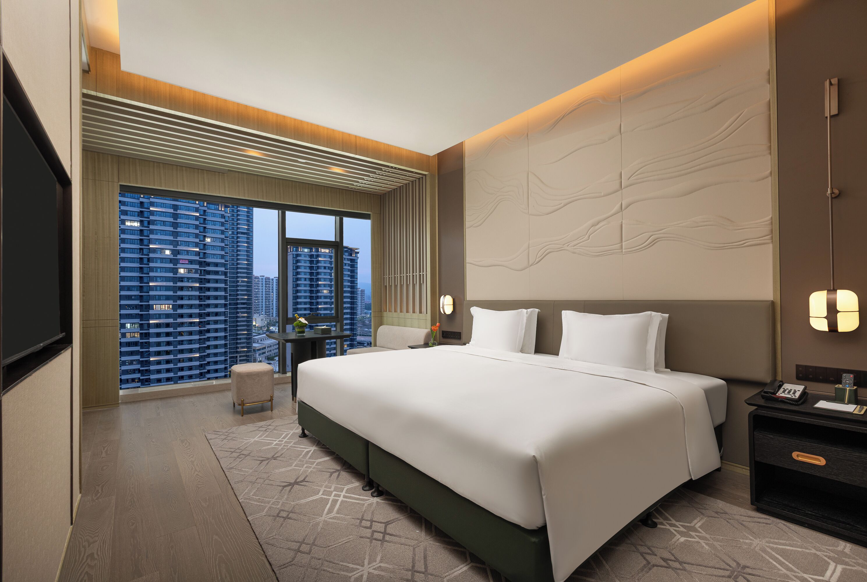 A modern hotel room with a large bed, a desk, and a view of tall buildings.
