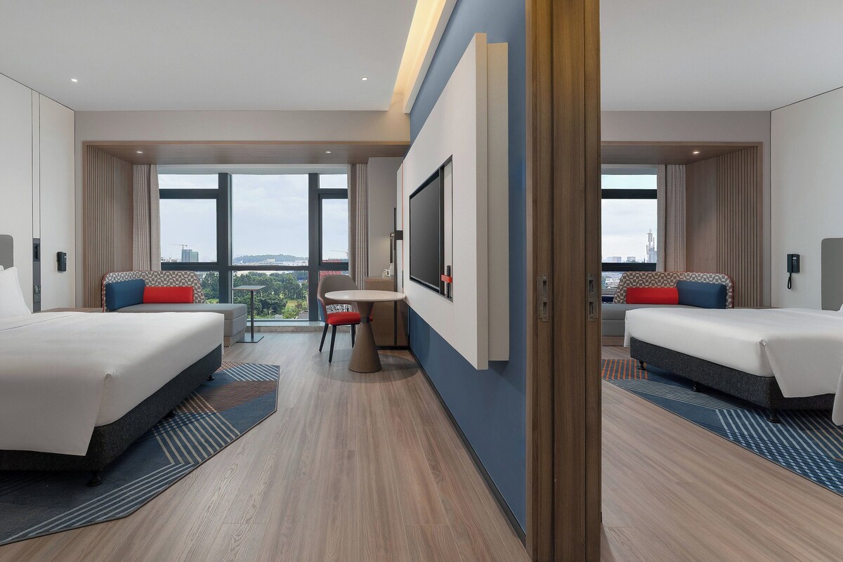A modern hotel room with two beds, a small table, and a large window offering a city view.