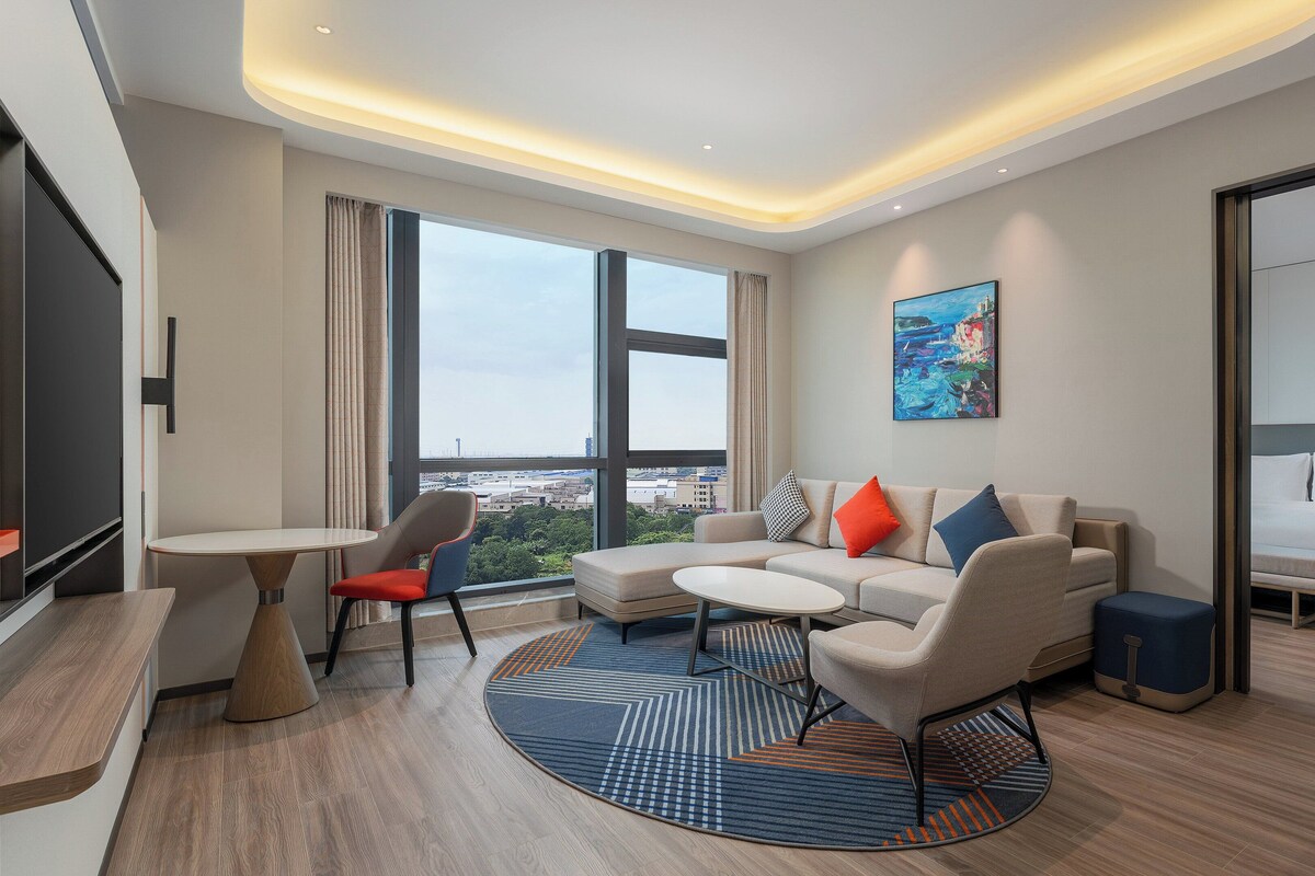 A modern hotel room with a large window, a sofa, a small table, and a bed.