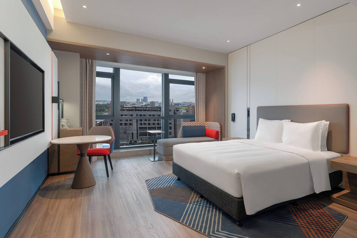A modern hotel room with a large bed, a desk, a chair, and a view of the city through large windows.